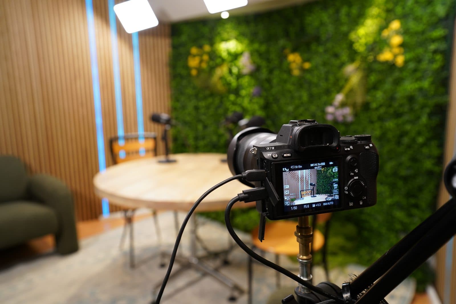 Sony-camera op statief klaar voor een opname, met de ronde tafel en groene plantenwand van de podcaststudio op de achtergrond