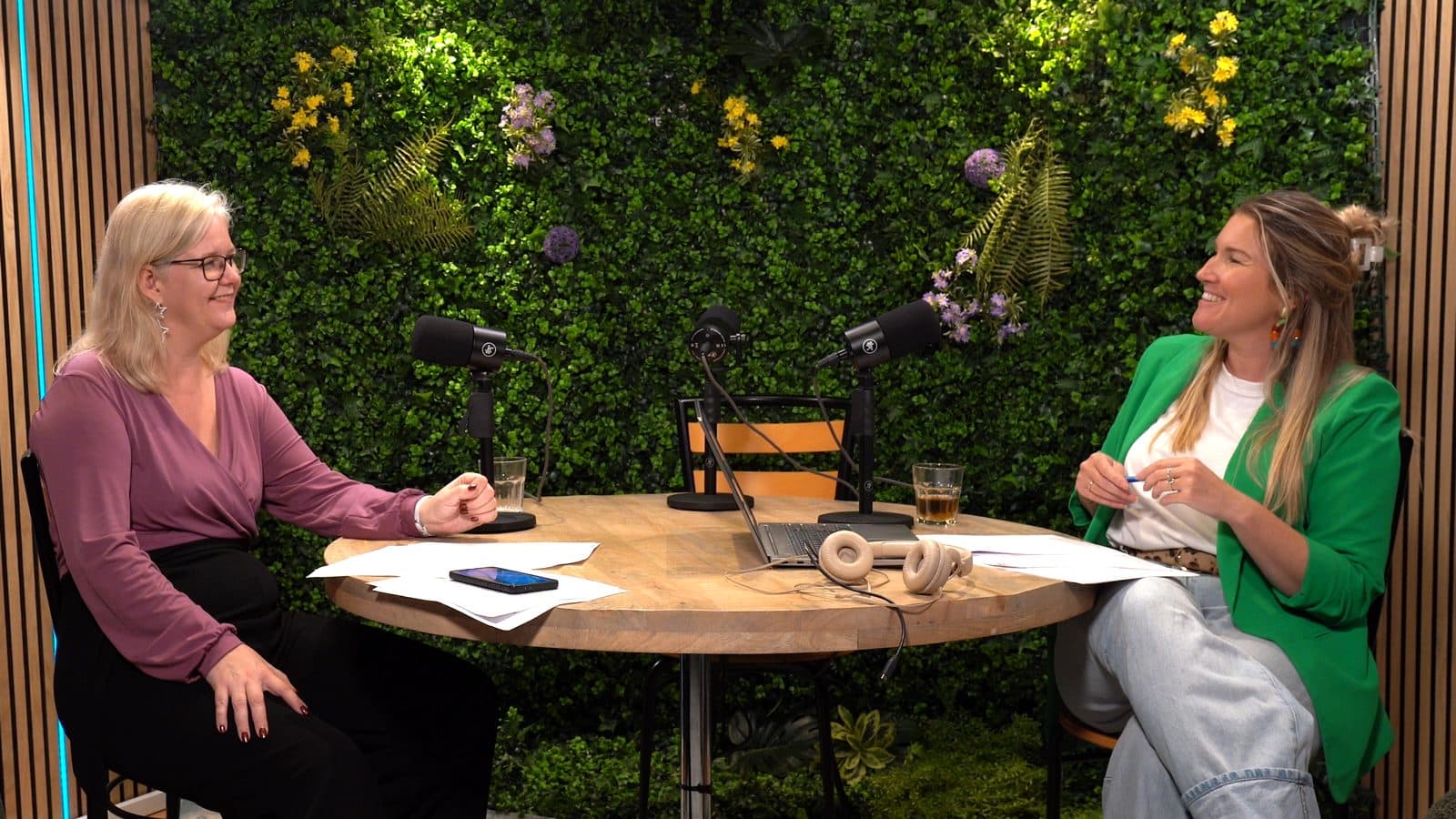 Twee vrouwen lachend in gesprek aan tafel tijdens een videopodcast-opname voor de groene plantenwand